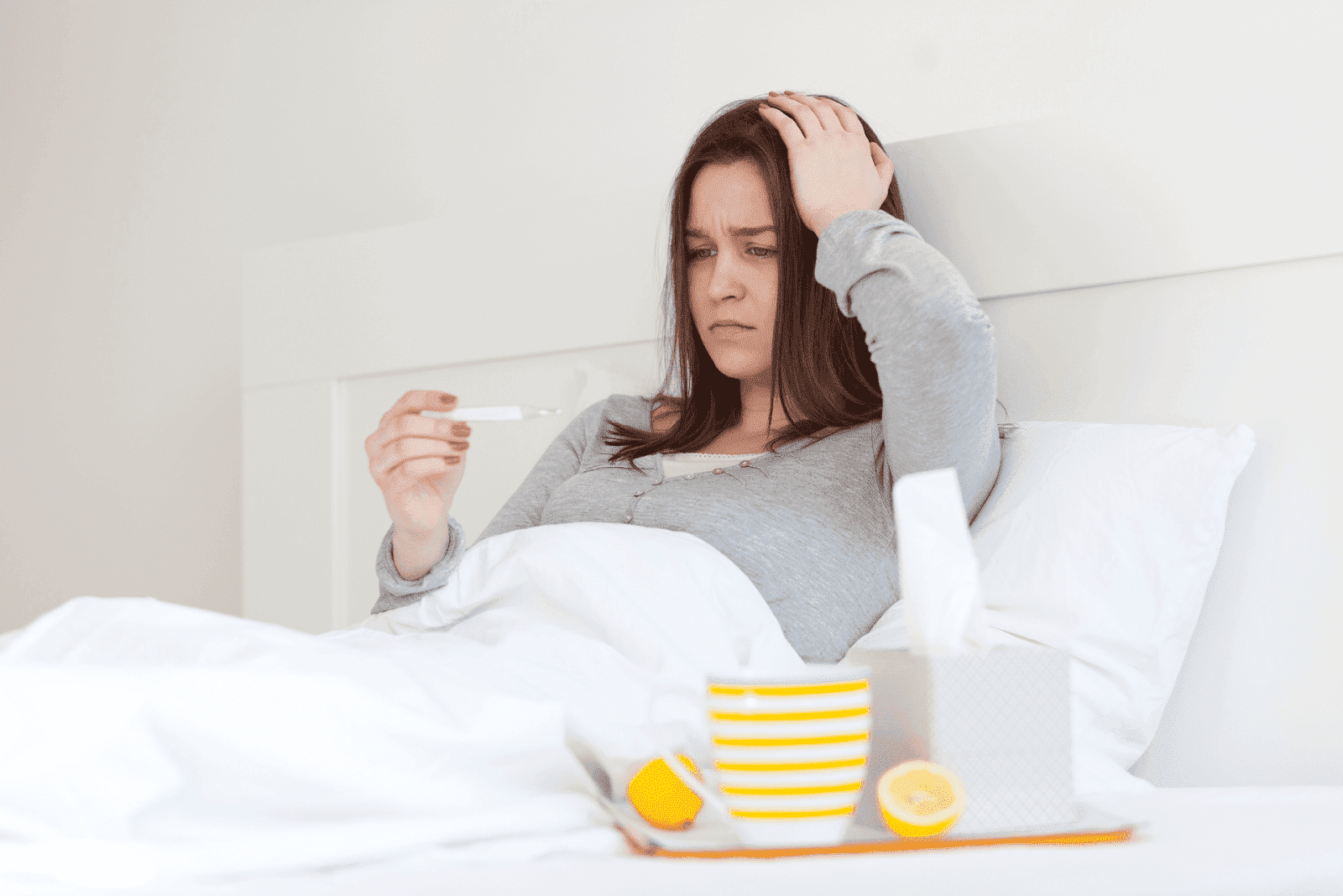 Woman in bed checking thermometer while looking unwell.