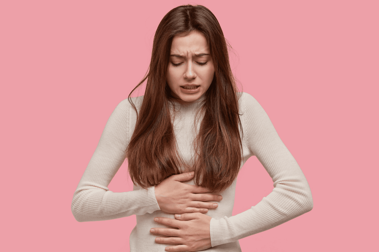Woman in discomfort clutching her stomach against a pink background.