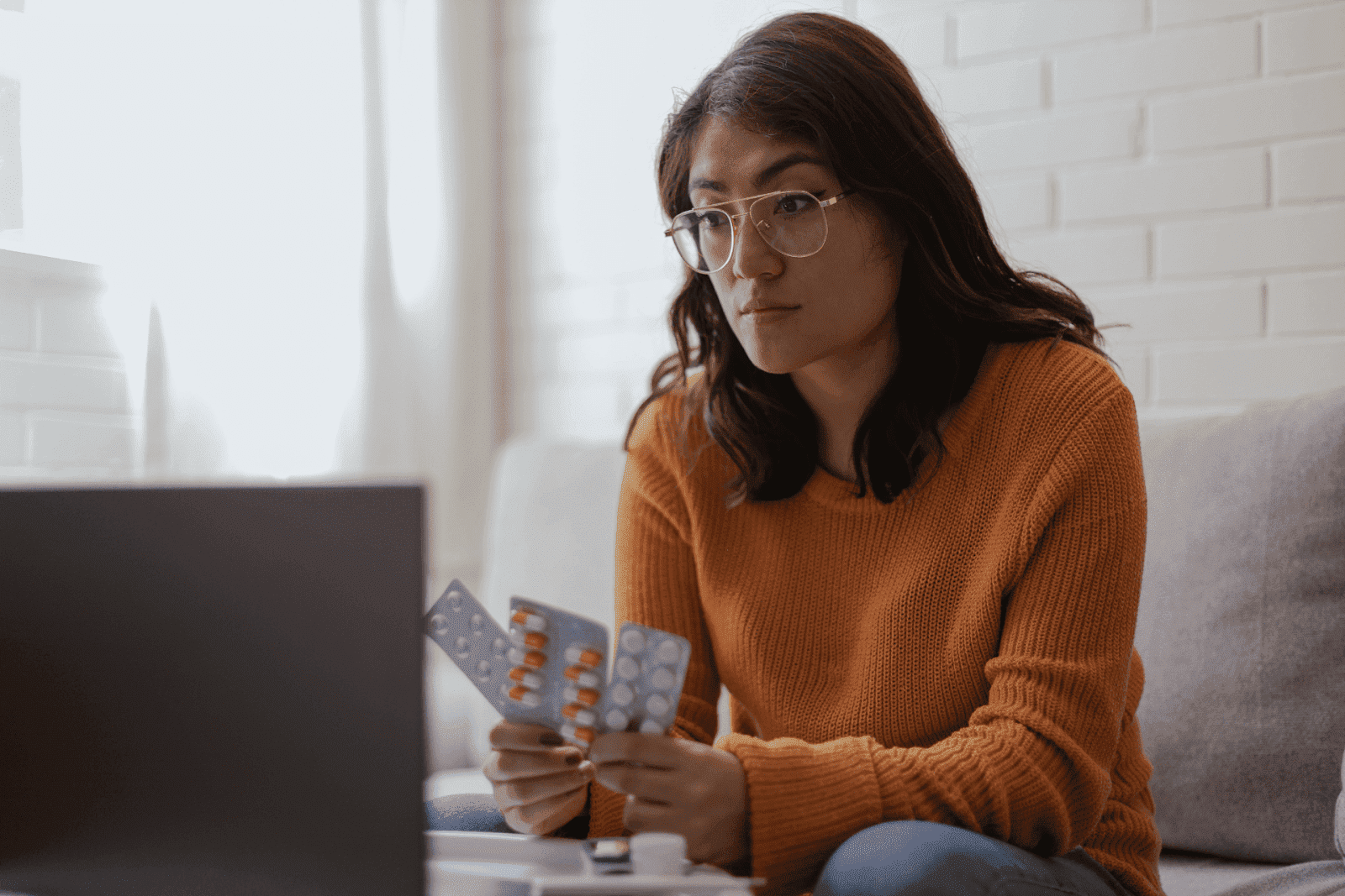 Woman in glasses holding medication blister packs while looking at a laptop.