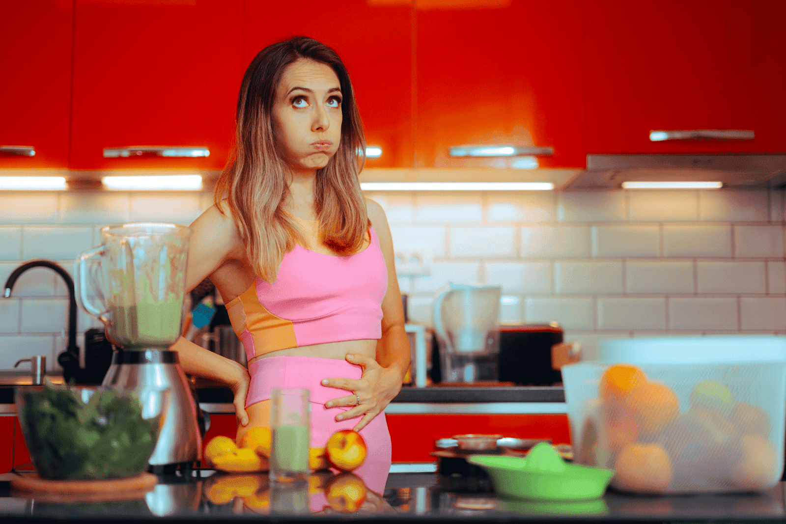 Woman in workout clothes holding her stomach and puffing cheeks in discomfort in a kitchen.