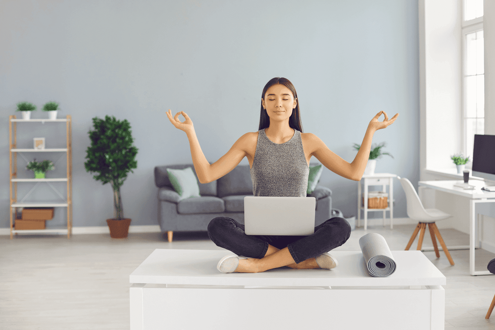 Woman meditating cross-legged on a desk with a laptop in front of her.