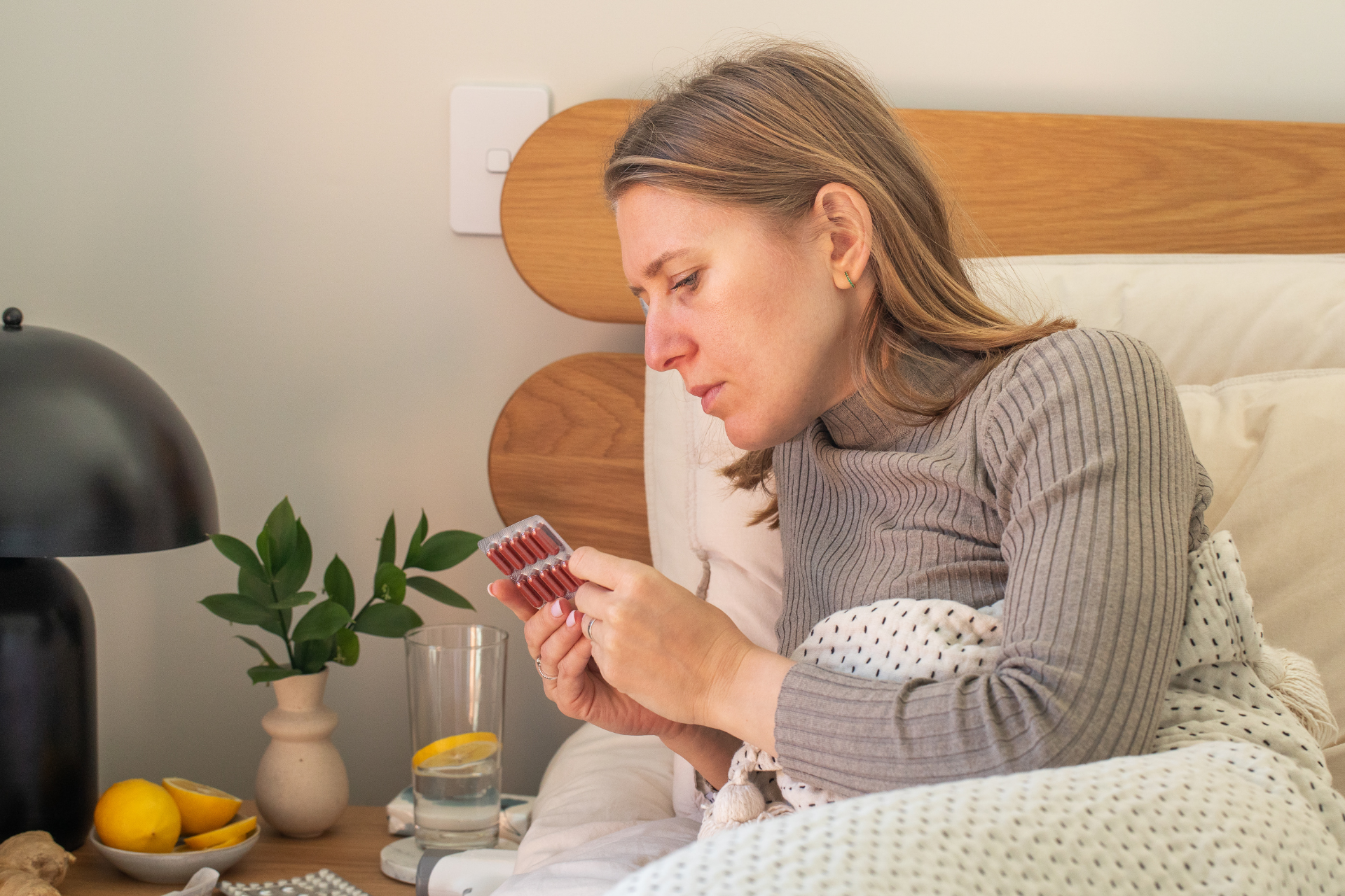Woman sitting in bed examining a blister pack of medication, appearing unwell