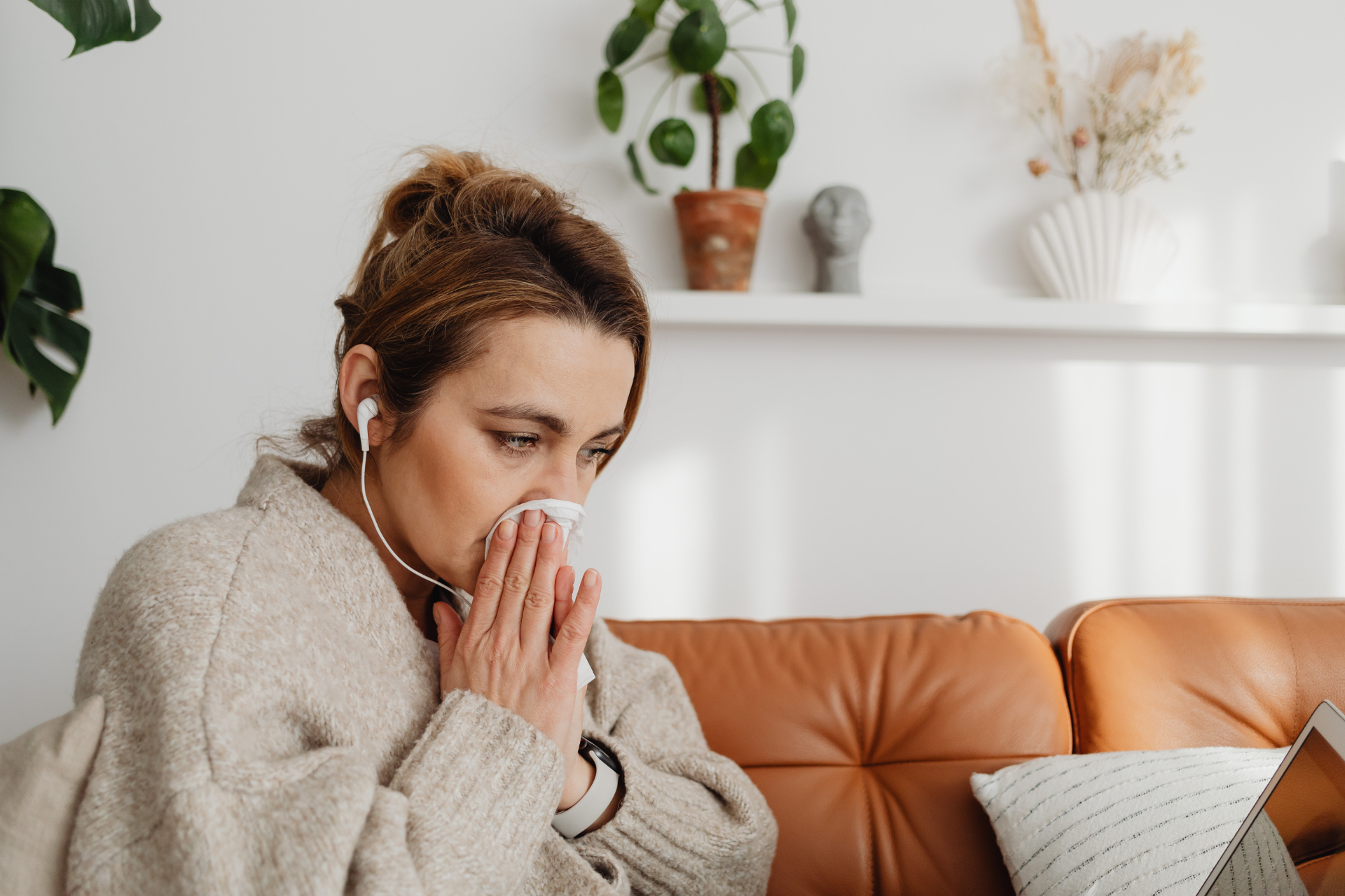 Woman sitting on a couch blowing her nose with a tissue, wearing earbuds and a sweater, appearing sick or congested in a home setting
