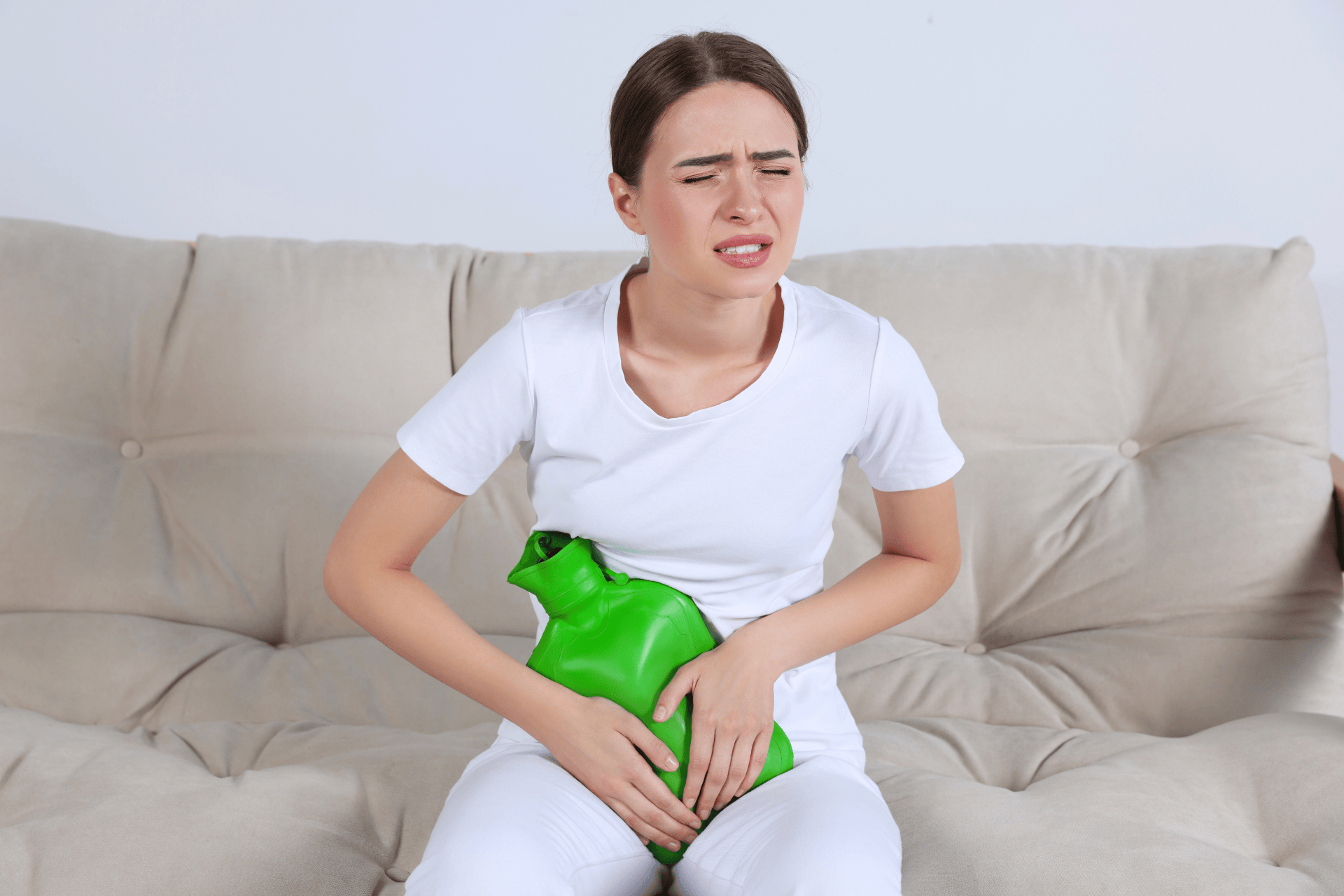 Woman sitting on a couch holding a green hot water bottle against her lower abdomen, appearing to be in pain.