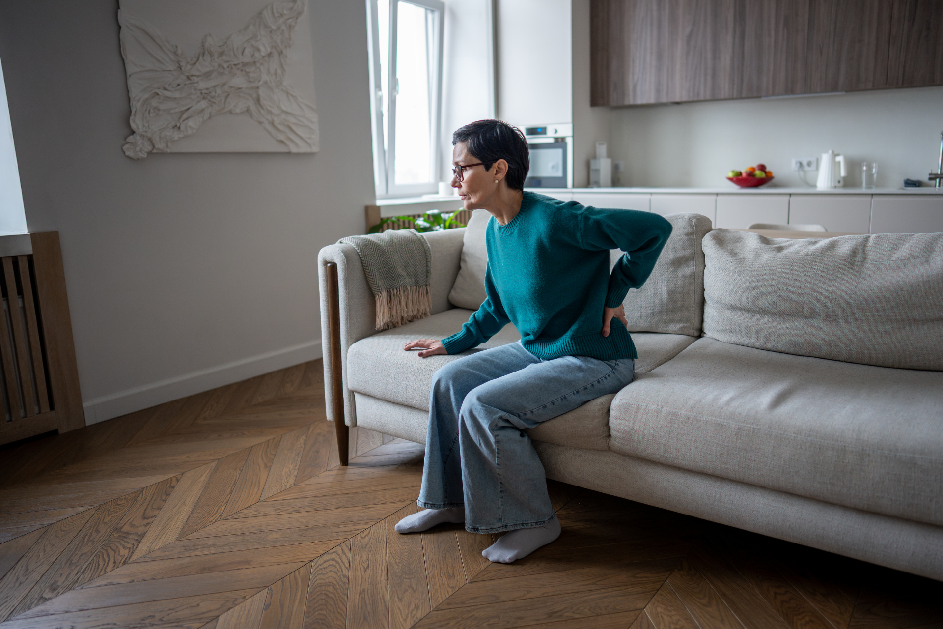 Woman sitting on a couch holding her lower back, showing back pain at home