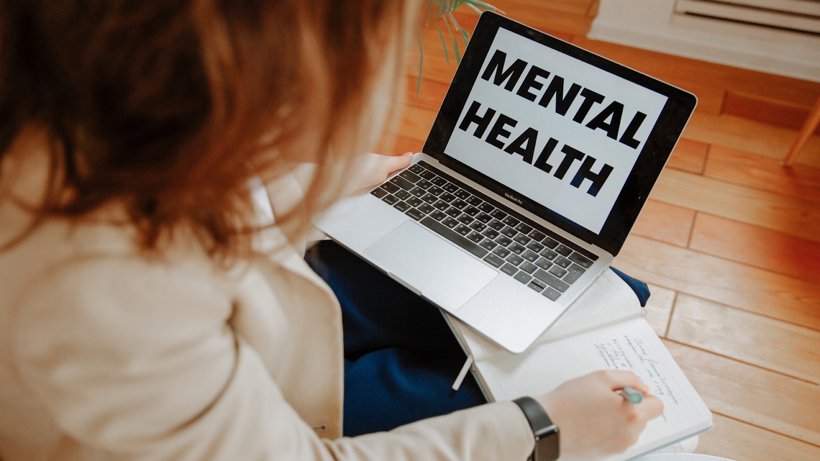 Woman watching a laptop screen that displays “MENTAL HEALTH” while taking notes in a notebook.