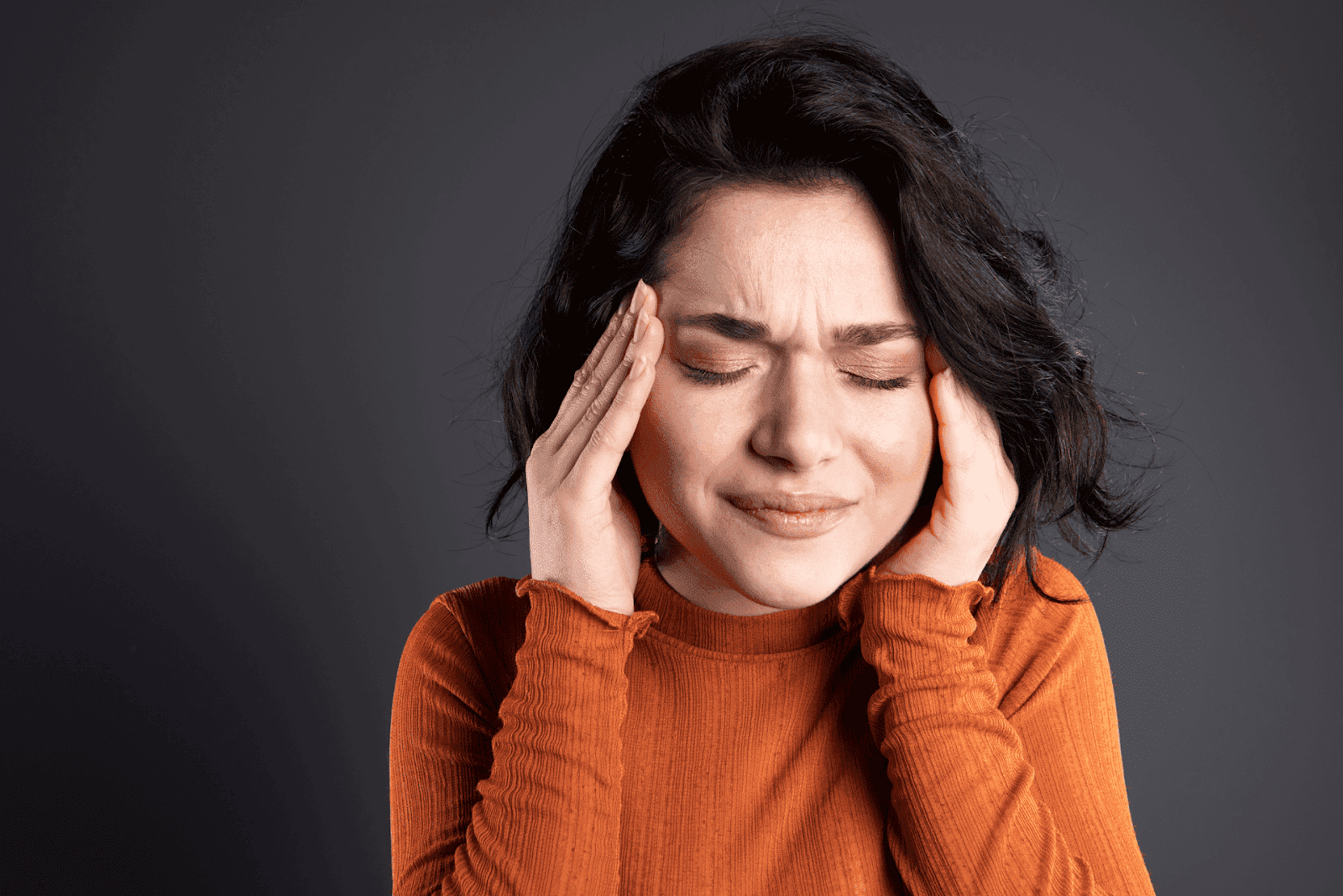 Woman with dark hair in an orange sweater holding her head in pain, showing signs of a headache or migraine