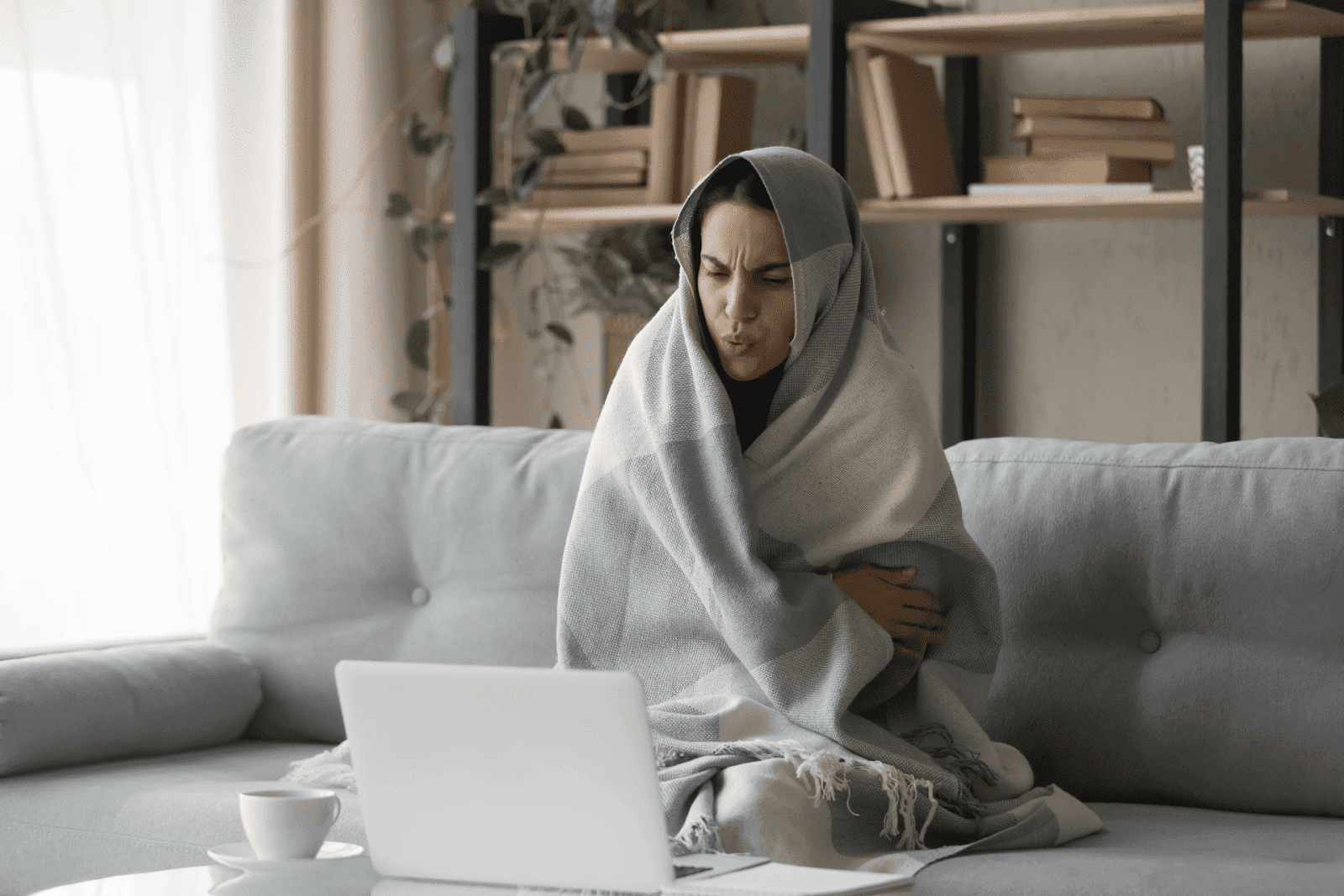 Woman wrapped in a blanket, shivering while sitting on a couch with a laptop
