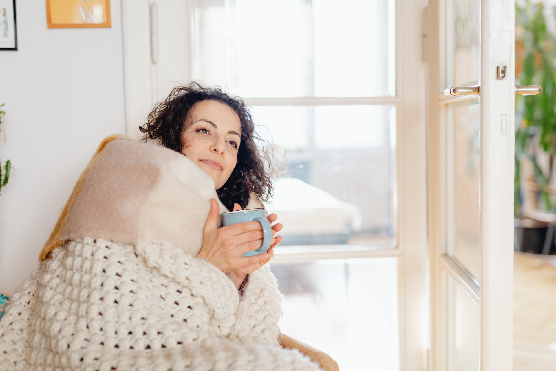 Woman wrapped in a blanket holding a warm mug by a window, appearing relaxed and recovering