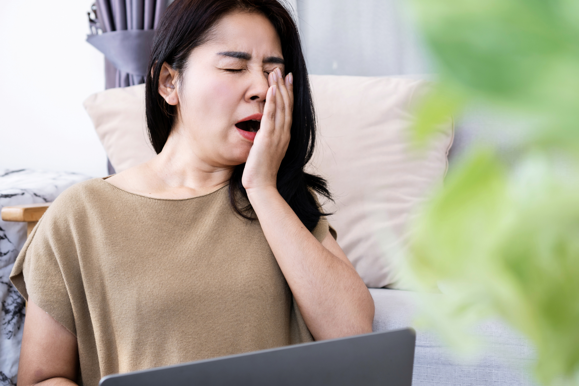Woman yawning and covering her mouth while sitting on a couch using a laptop, appearing tired or fatigued