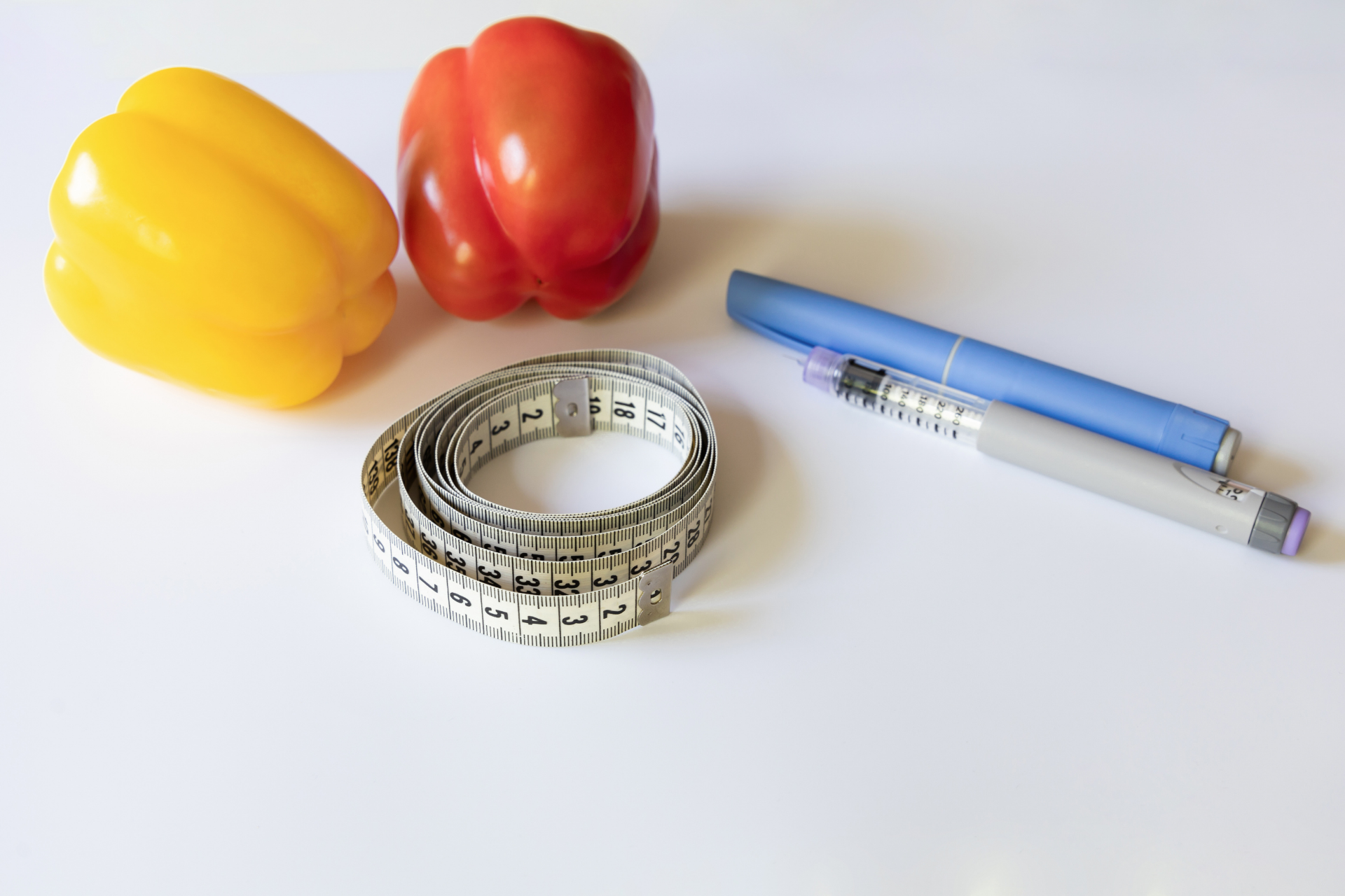 Yellow and red bell peppers, a coiled measuring tape, and an injectable medication pen on a white background, representing weight management, nutrition, and diabetes treatment