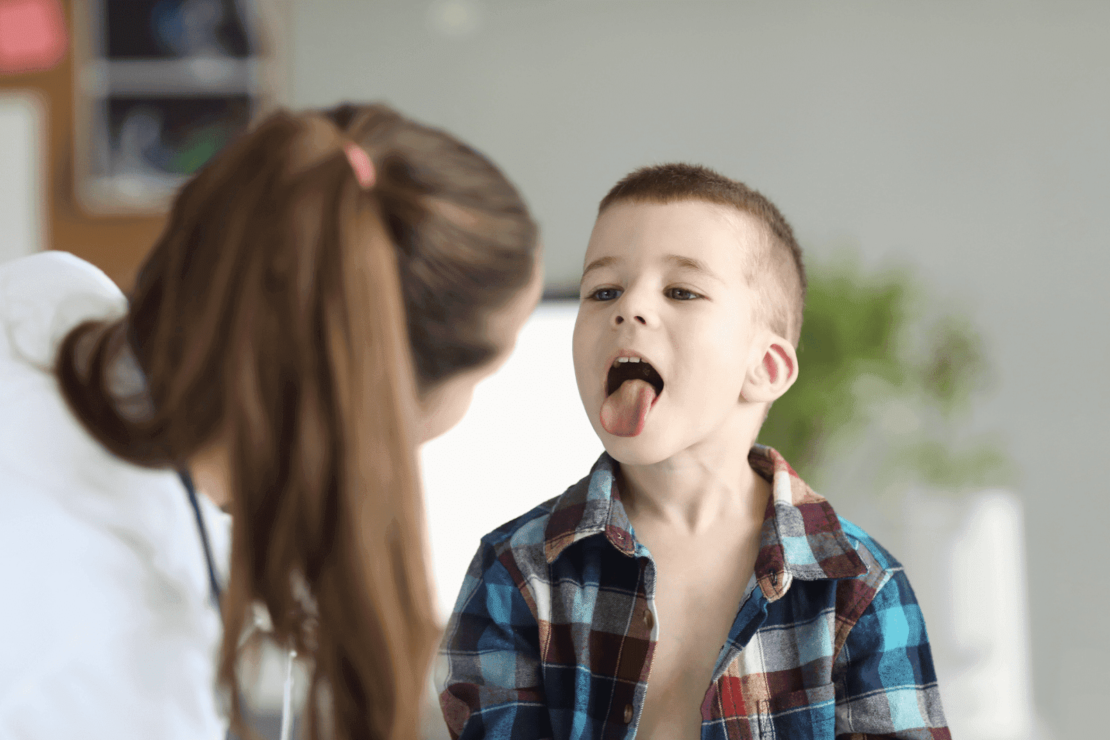 Young boy in a plaid shirt sitting with his mouth open and tongue out while a healthcare professional observes him during a medical checkup