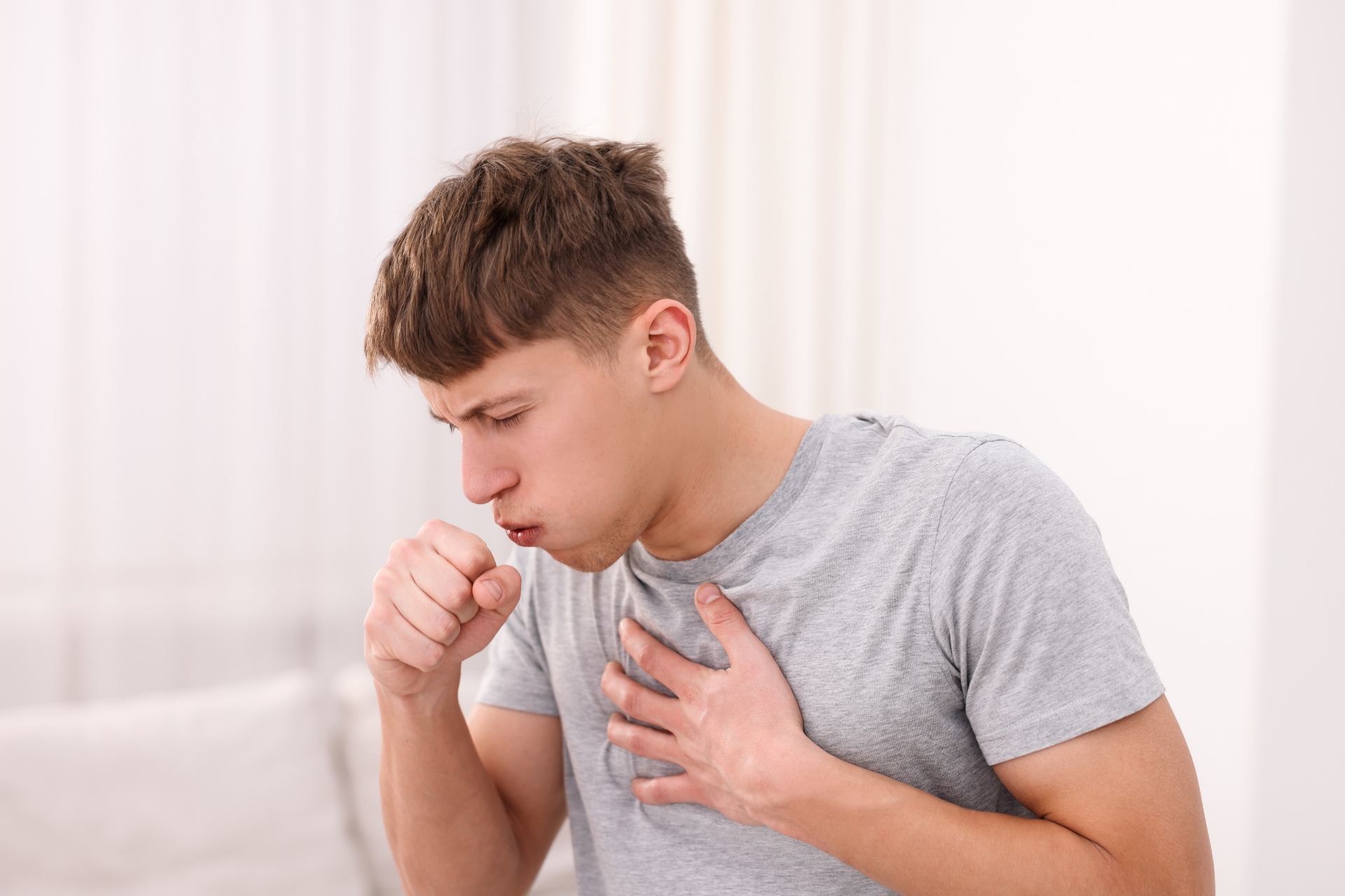 Young man coughing into his fist while holding his chest, appearing uncomfortable or experiencing a persistent cough